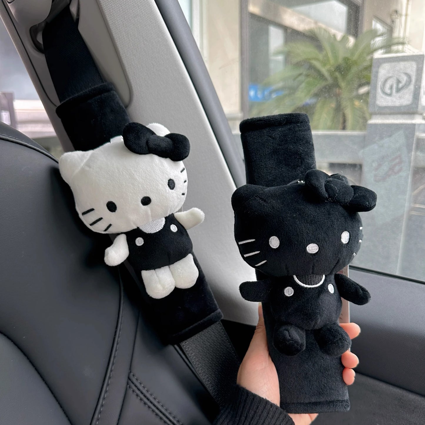 Two plush Hello Kitty car seatbelt covers in black and white, held by a hand inside a car.