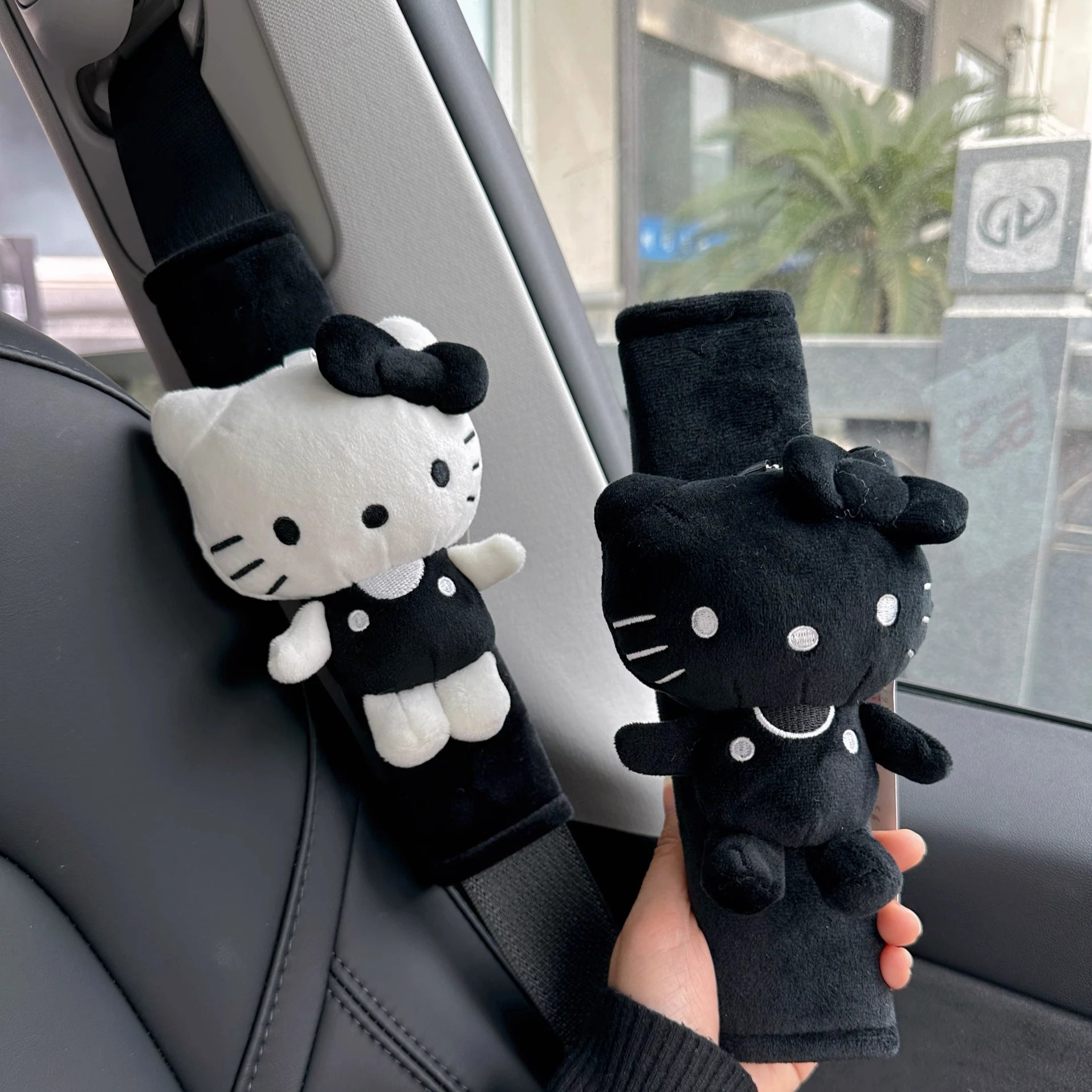 Two plush Hello Kitty car seatbelt covers in black and white, held by a hand inside a car.